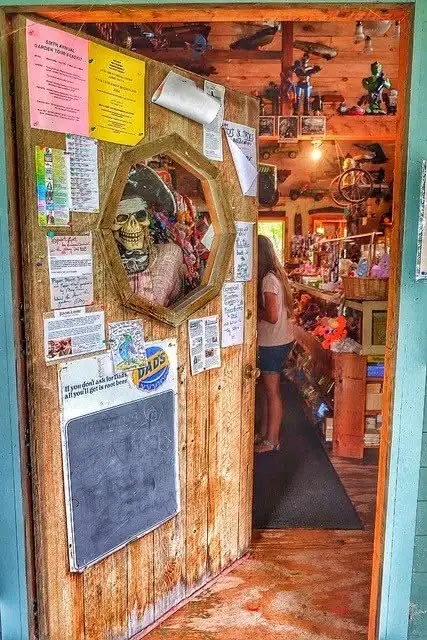 A weathered wooden shop door displays a framed skull decoration, posted notices, and a chalkboard beside an open storefront.