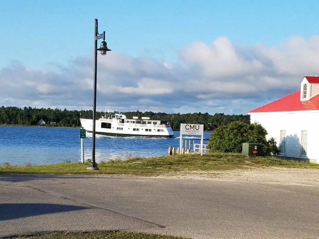 A passenger ferry passes a waterfront road beside a red-roofed building and a university sign.