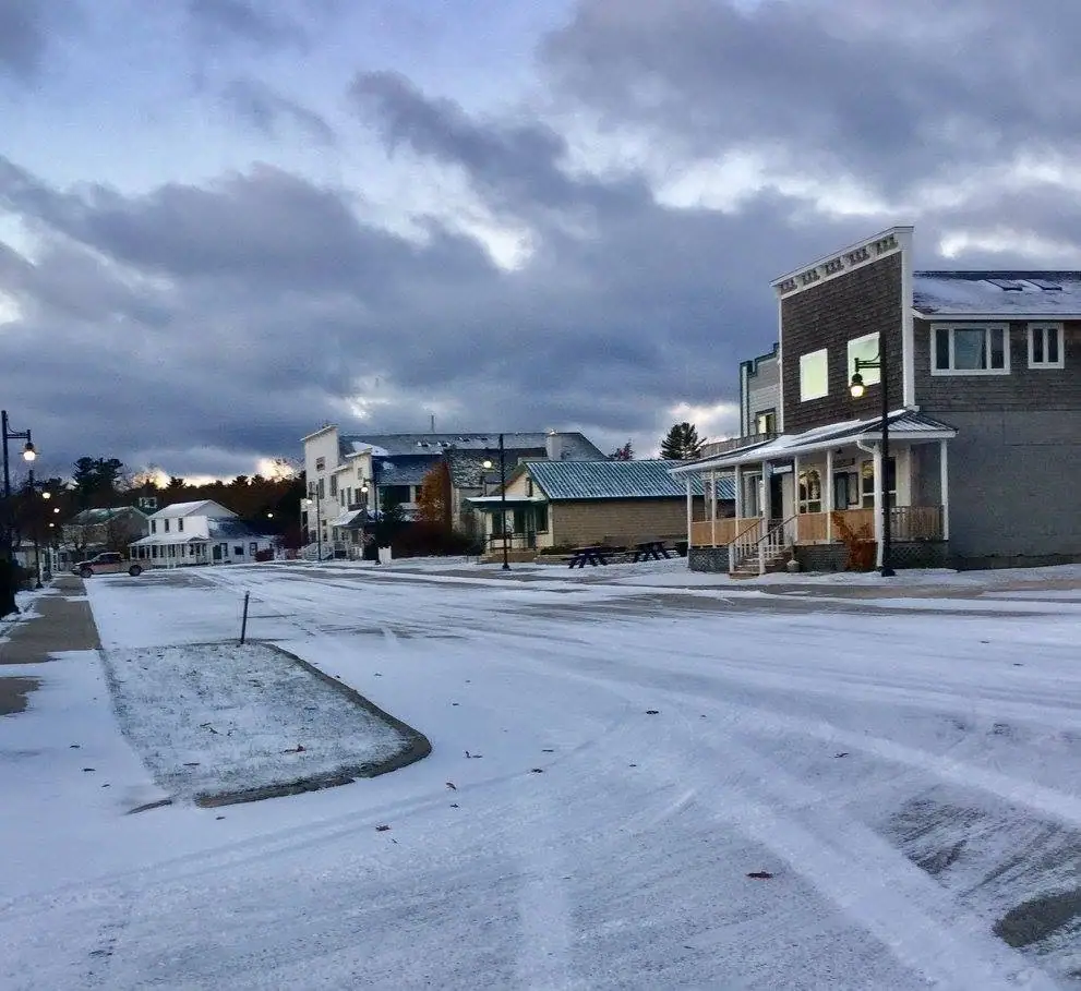 Snow-covered town street lined with storefronts and houses beneath a cloudy evening sky.