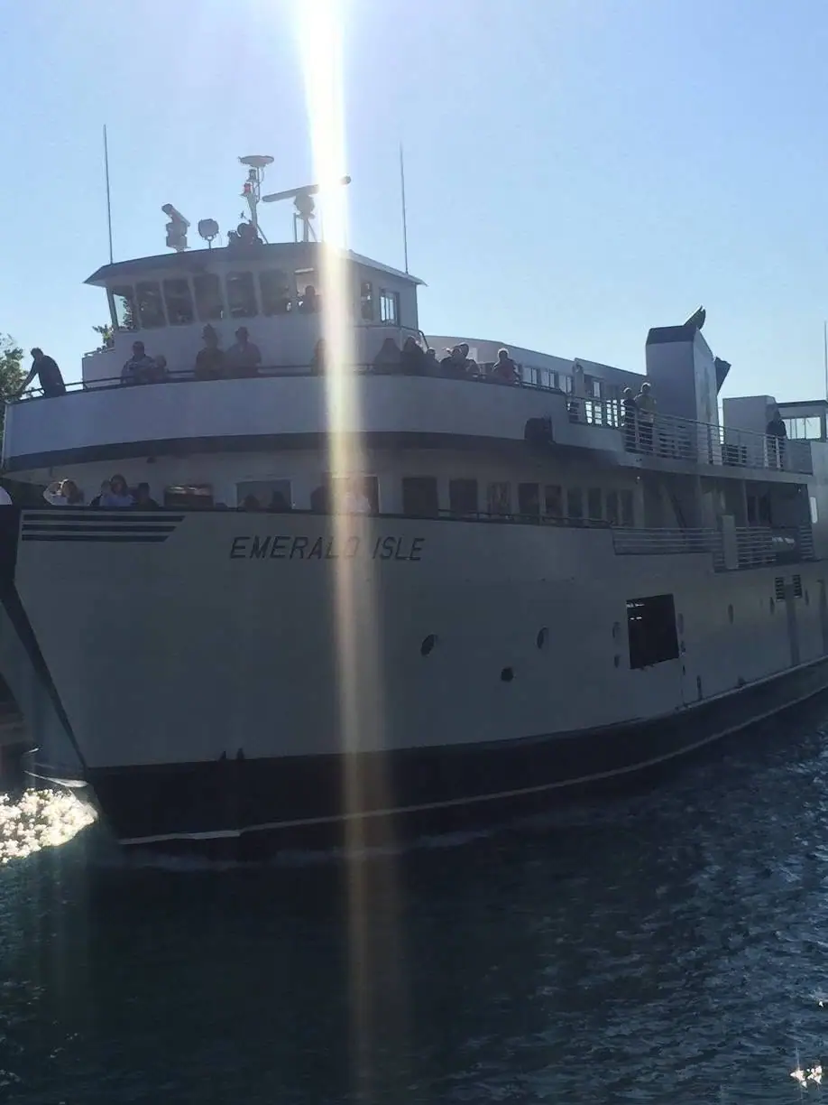 A large white passenger ferry carrying people moves through dark, rippling water in bright sunlight.
