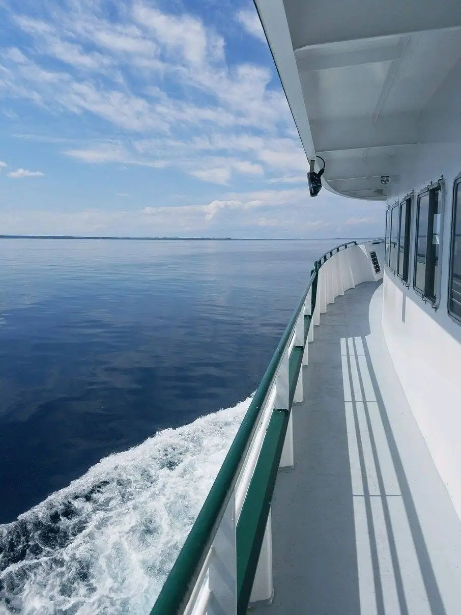 A ferry’s side deck and green railing overlook its foaming wake and calm open water beneath a partly cloudy sky.