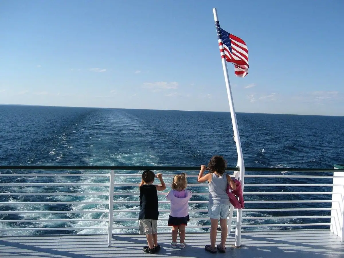Three children stand at a ferry railing beneath an American flag, watching the vessel’s wake across open water.