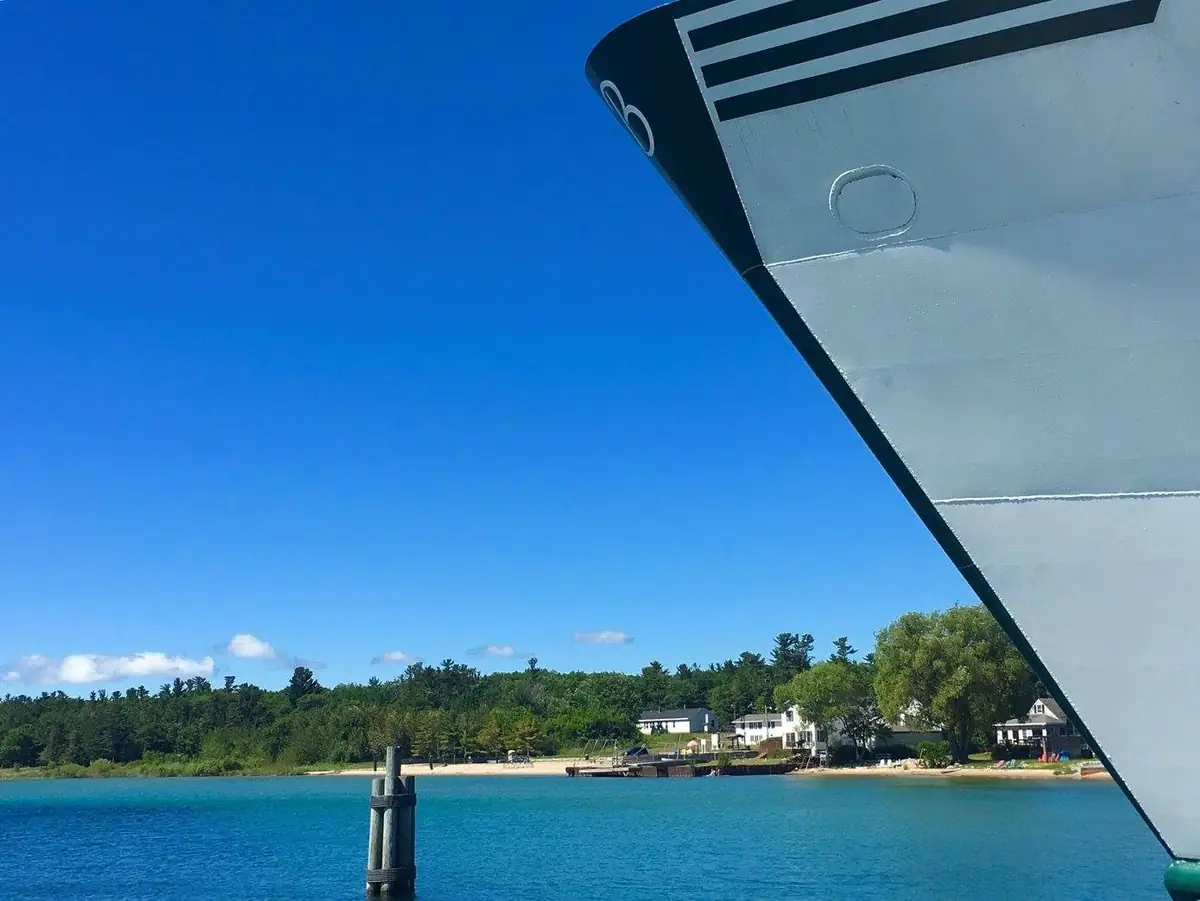 A ferry's white bow frames a wooded shoreline, beach, dock, and waterfront houses.