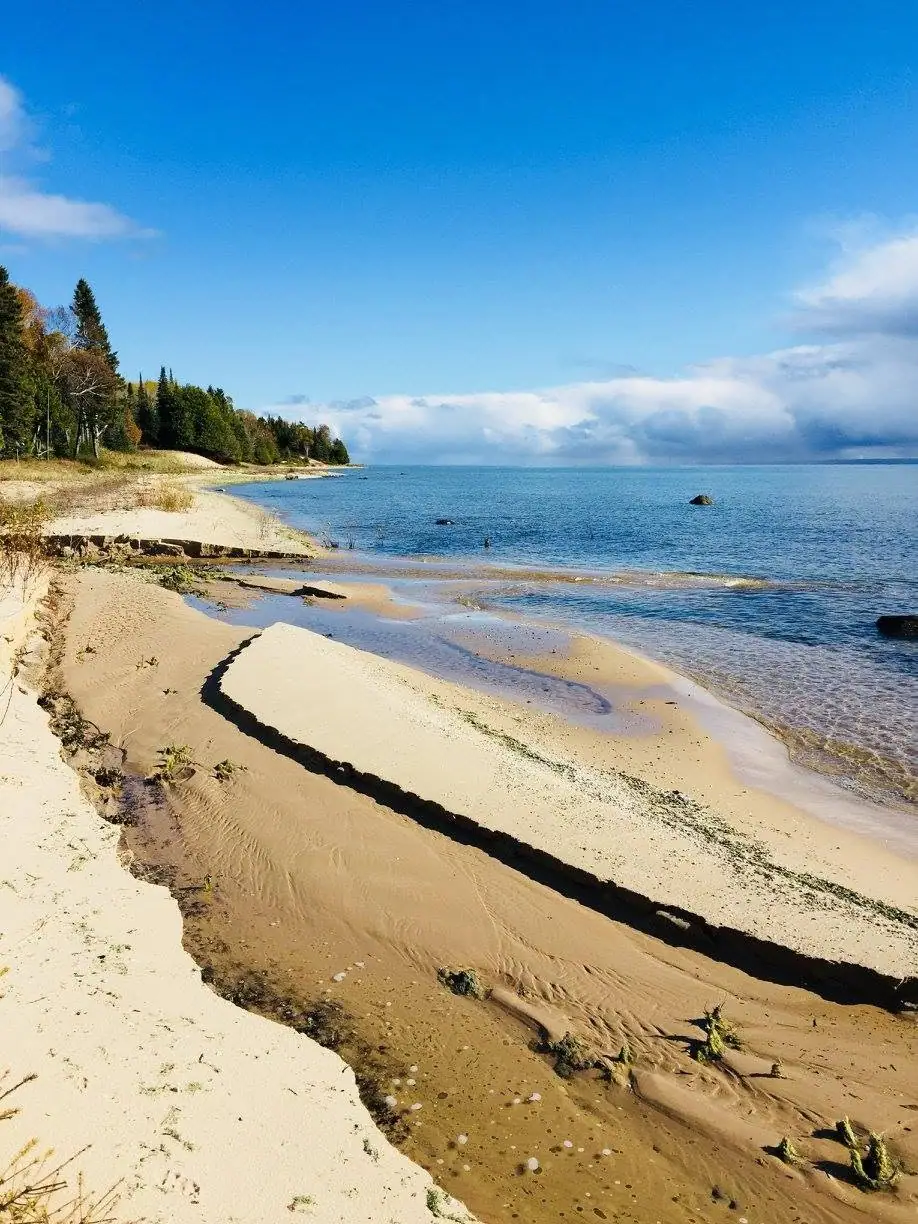 A sandy shoreline with a narrow stream, forest, and blue water beneath a partly cloudy sky.