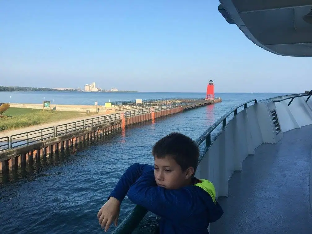 A child in a bright vest leans on a ferry railing beside a pier and red lighthouse.