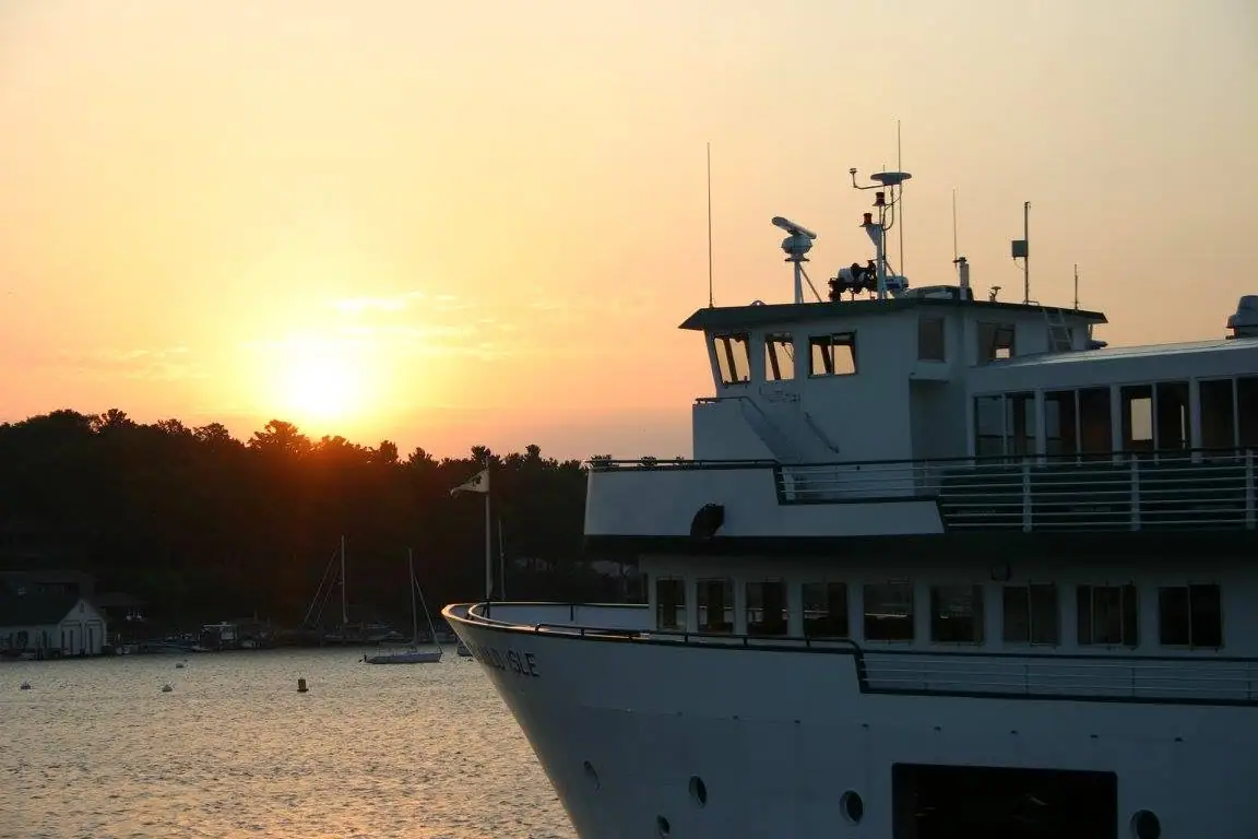 A large white ferry passes through sunset-lit water near smaller sailboats and a tree-lined shore.