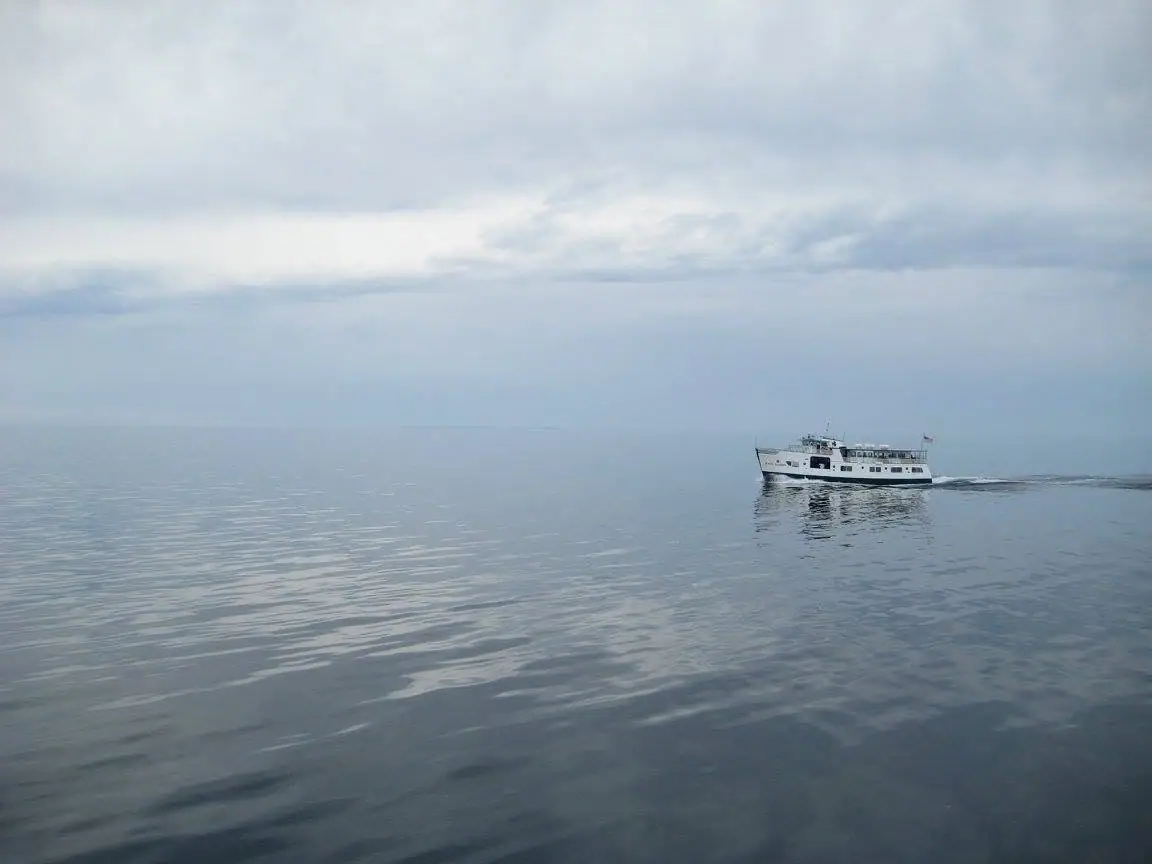 A white passenger ferry moves across calm water beneath a broad, cloudy sky.
