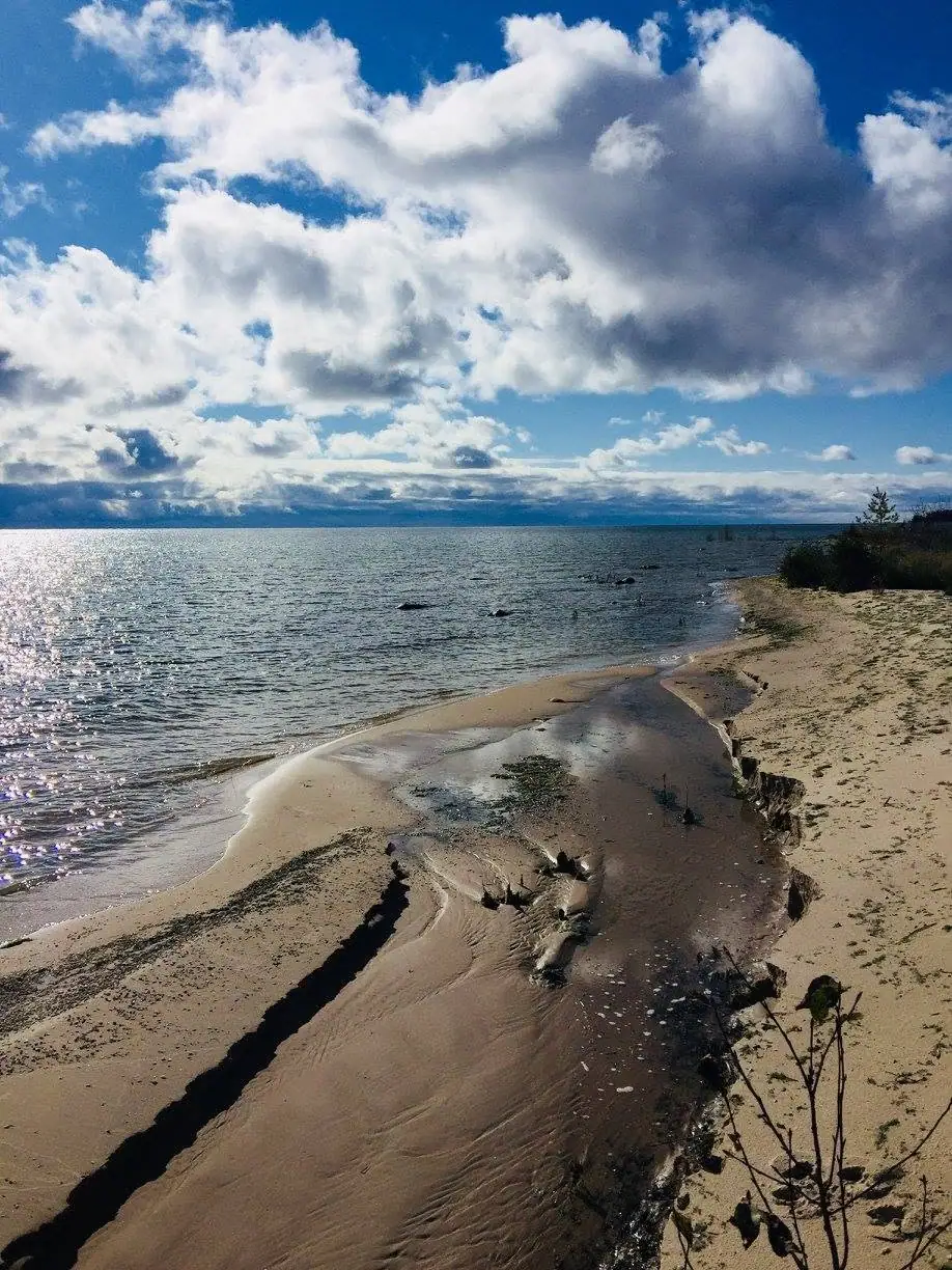 A sandy shoreline curves beside rippling water beneath towering clouds, with a shallow channel cutting through the beach.