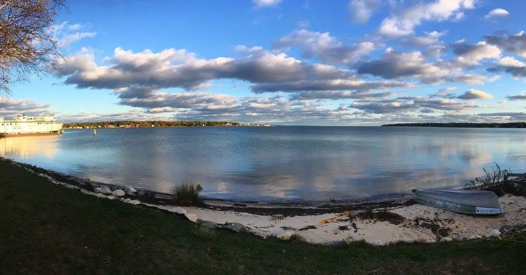 A white ferry and small boat sit beside a sandy shoreline beneath a cloud-filled blue sky.