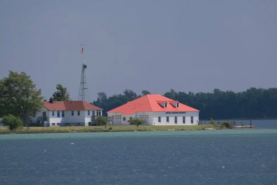 Two white, red-roofed buildings stand beside the water, with a tower and flag behind them.
