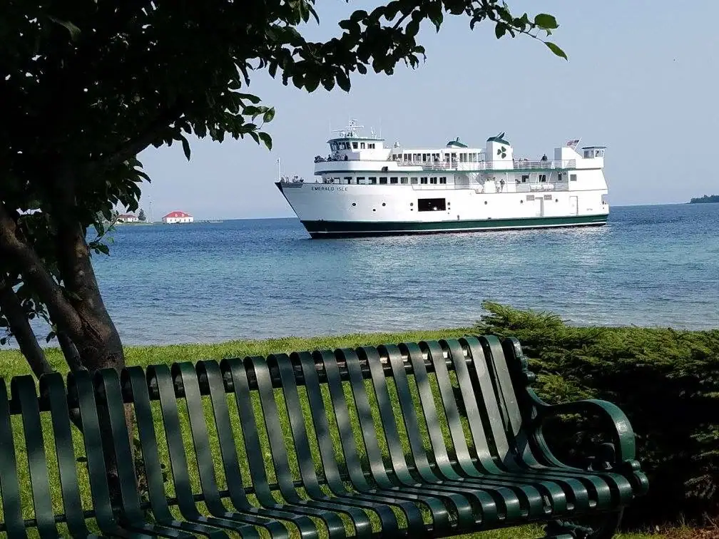 A white passenger ferry passes offshore, seen beyond a green park bench and leafy tree.