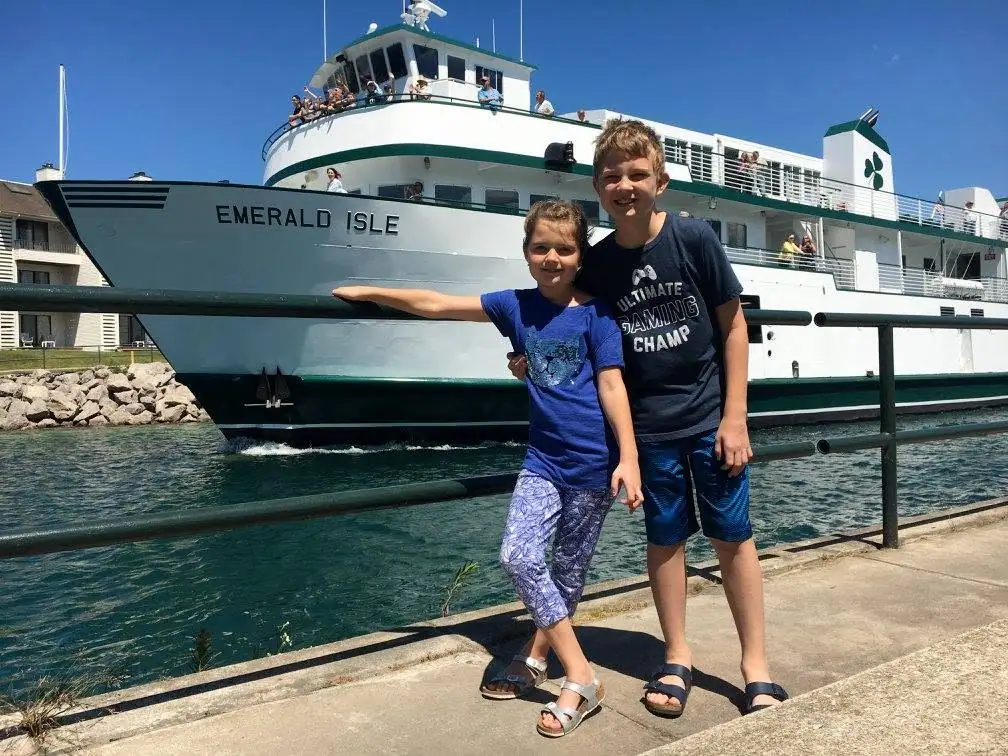 Two children pose beside a white-and-green passenger ferry at a waterfront dock.