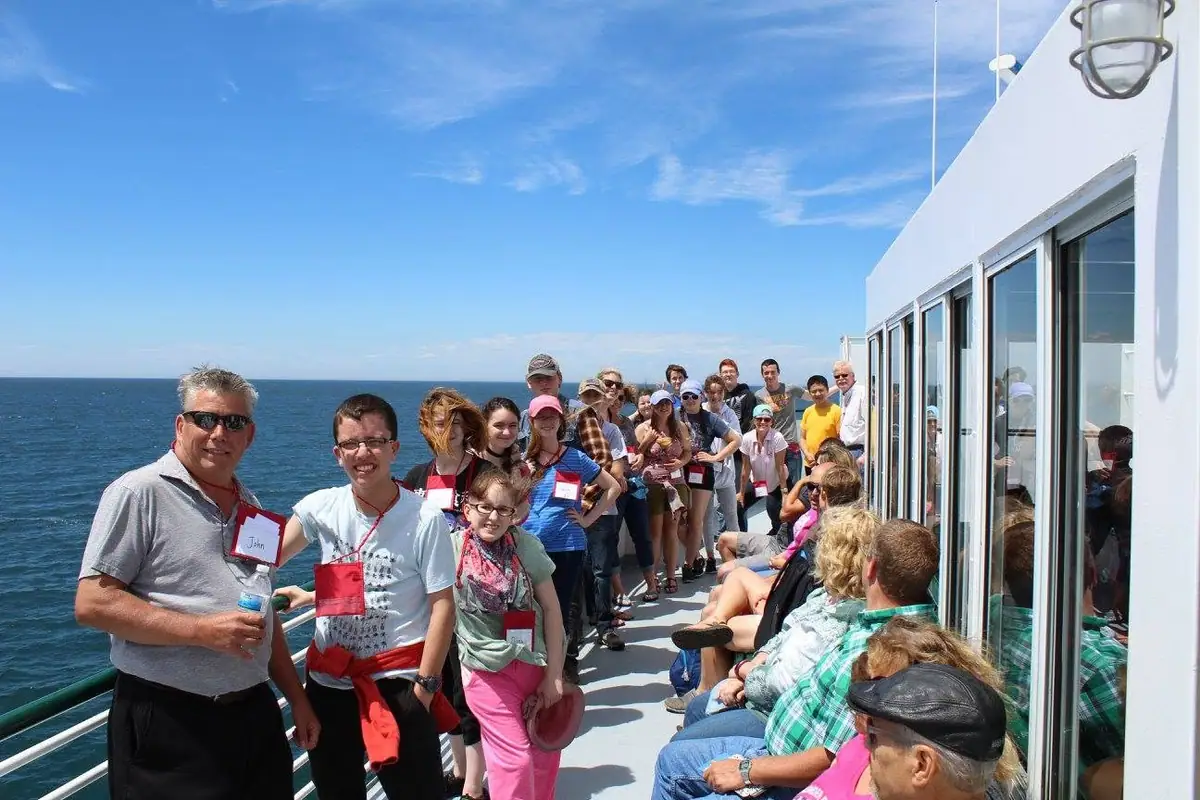 A group of passengers gathers on the sunny deck of a ferry beside open water.