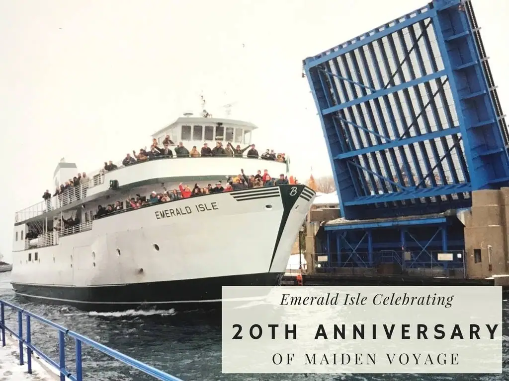 A crowded white passenger ferry passes beneath a raised blue drawbridge beside a waterfront railing.