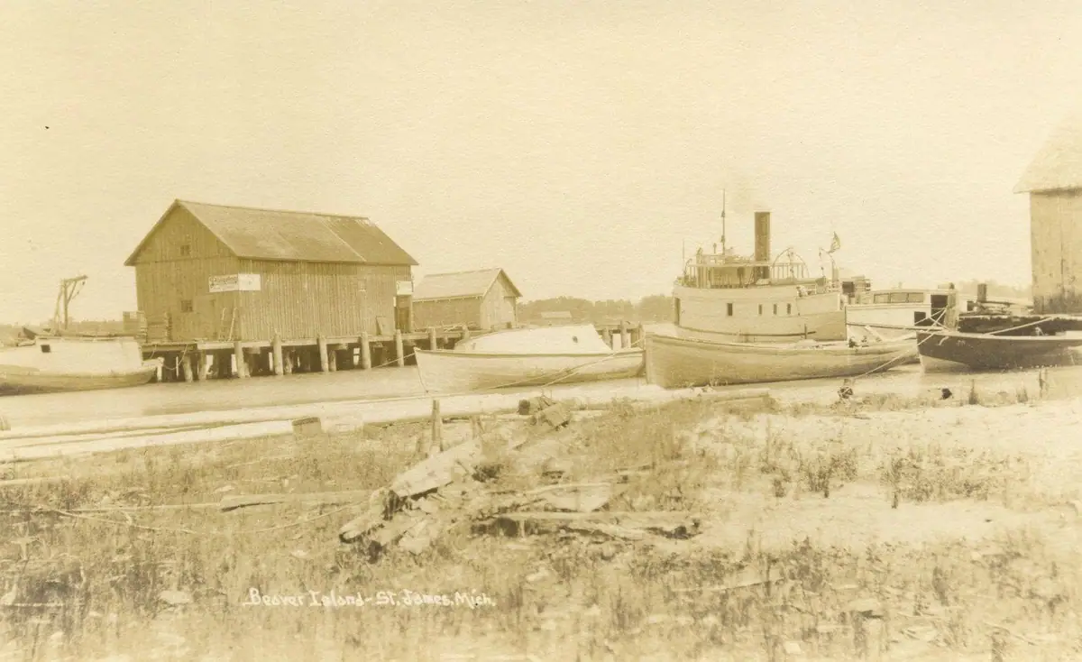 Sepia harbor photograph with waterfront buildings, a steamer, smaller boats, and a cylindrical storage tower.