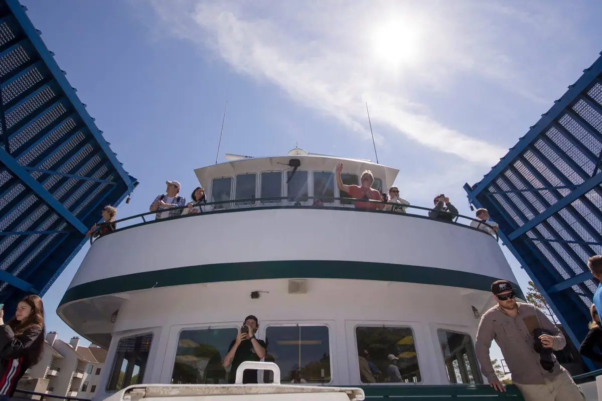 Passengers wave from the upper deck of a white ferry framed by blue steel structures.