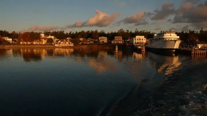 A white ferry sits beside a pier across calm water, reflecting shoreline houses and trees in warm evening light.