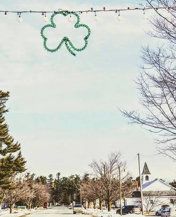 Winter street scene with a shamrock-shaped decoration, overhead lights, cars, and a white steepled church.