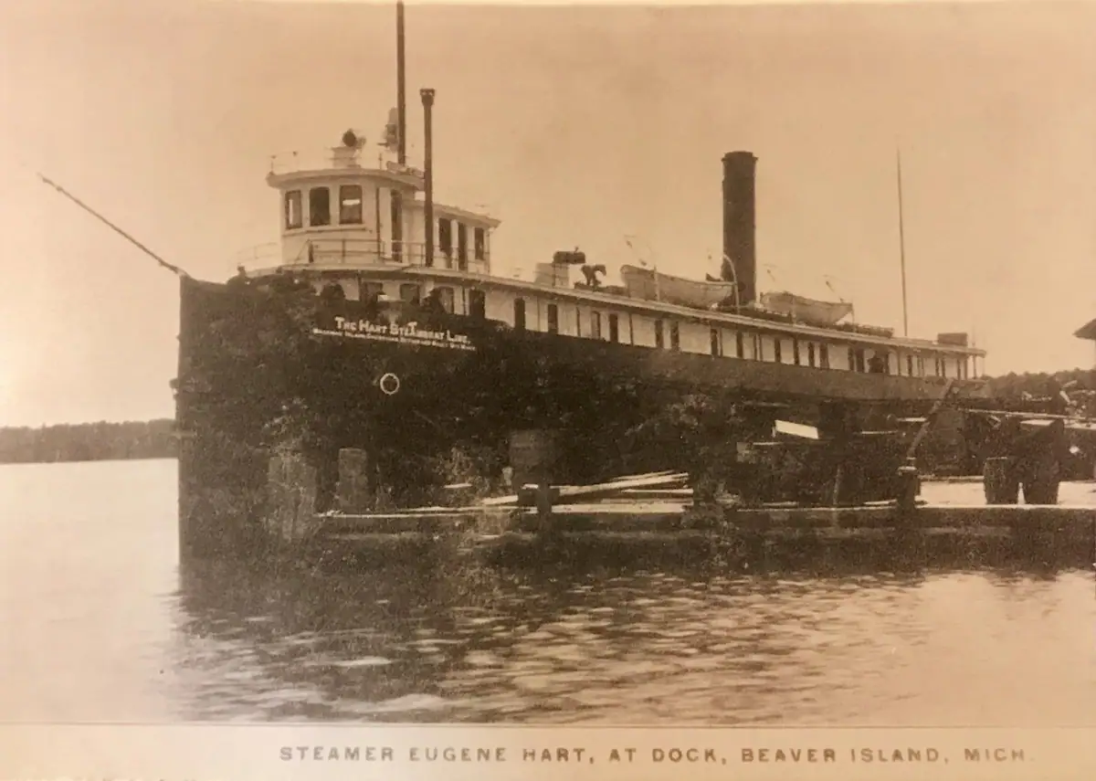 Sepia photograph of a large steamer moored beside a timber pier, with a tree-lined shore in the background.