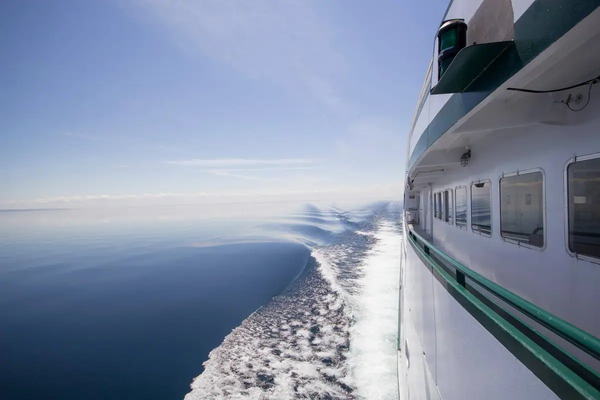 A white passenger ferry moves across calm water, leaving a foaming wake beneath a clear sky.
