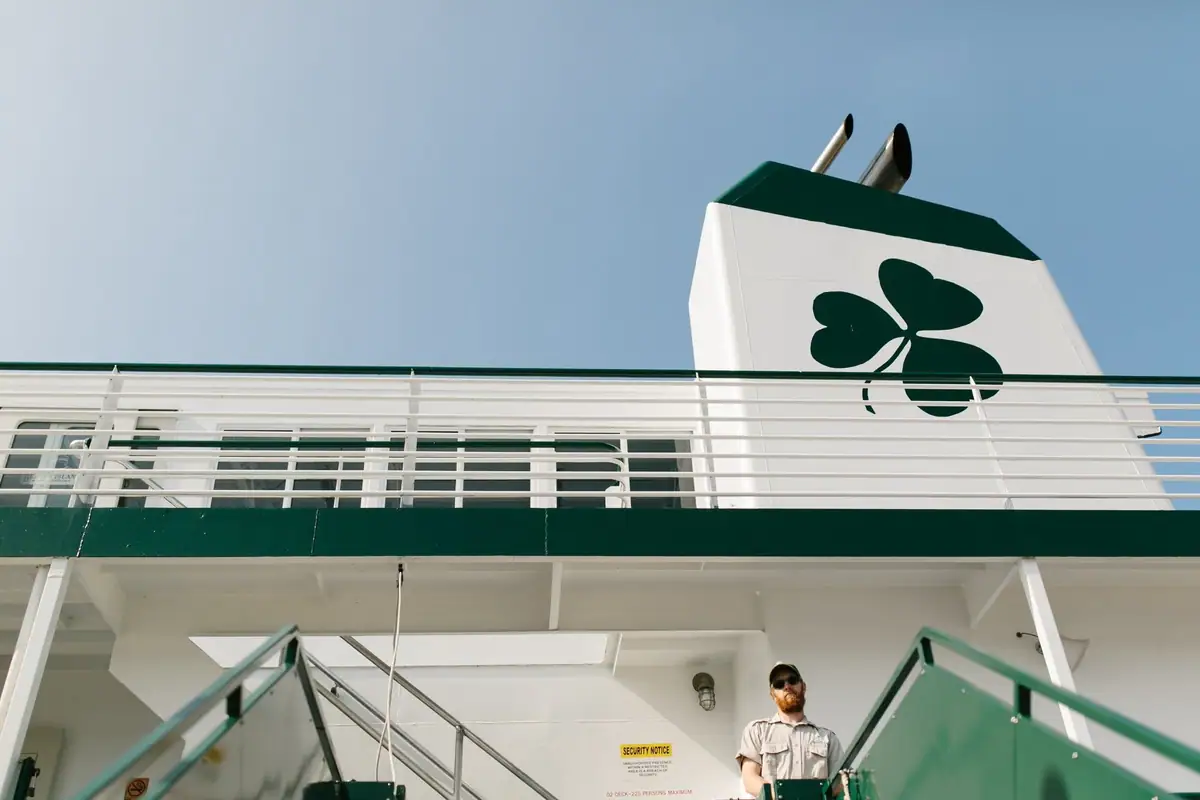 Low-angle view of a ferry deck with a large green shamrock, exhaust pipes, railings, and a uniformed man.