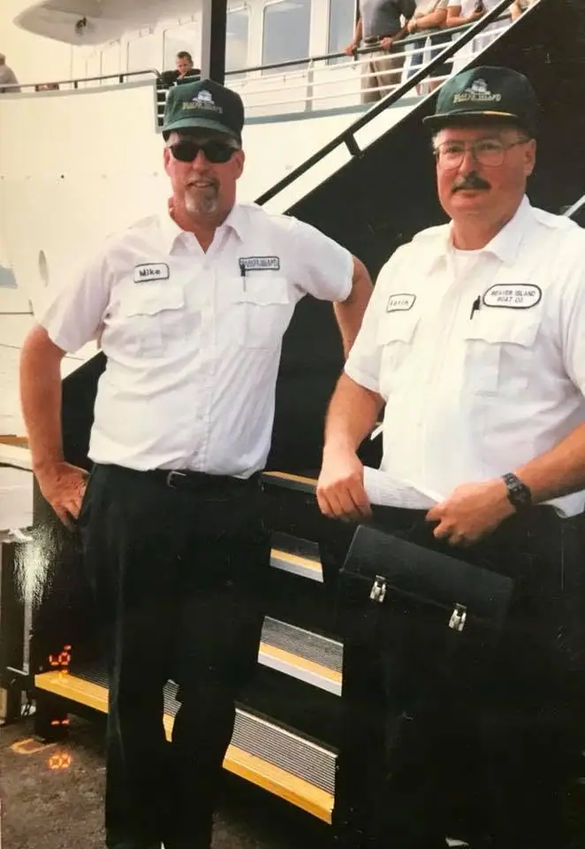 Two men in white crew uniforms and green caps stand on a vessel deck beside a stairway.