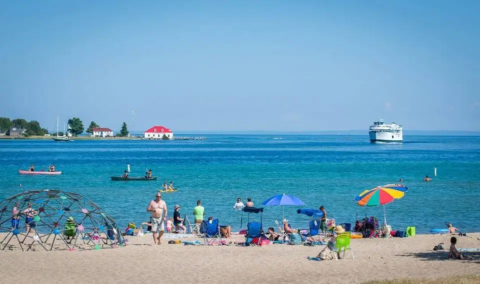 People relax and play along a sunny sandy beach while a ferry crosses the water beyond them.