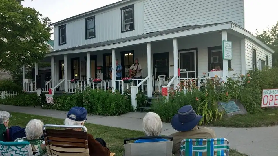 Two musicians perform on a porch while seated listeners watch outside a white two-story building.