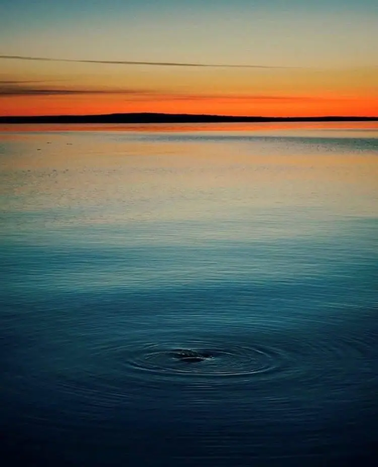 Warm orange light spreads across calm water toward a dark distant shoreline.