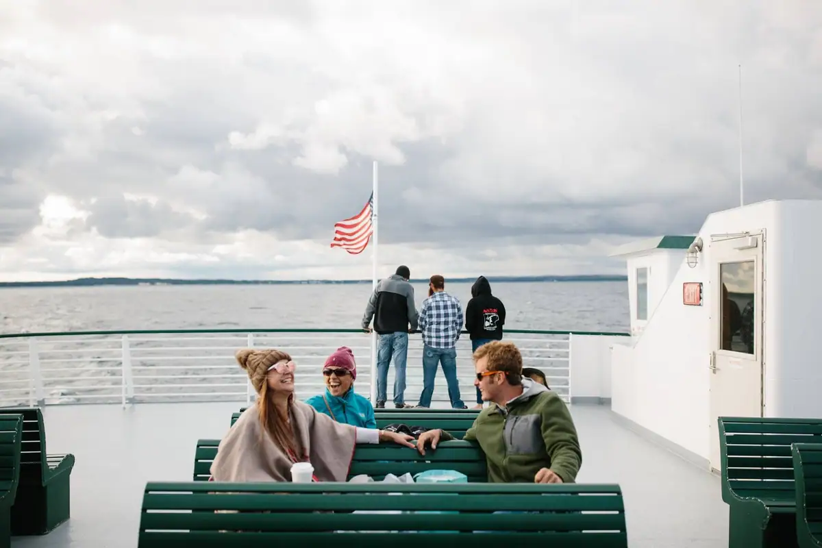 Adults smile and stand on the open deck of a ferry beneath a cloudy sky and an American flag.