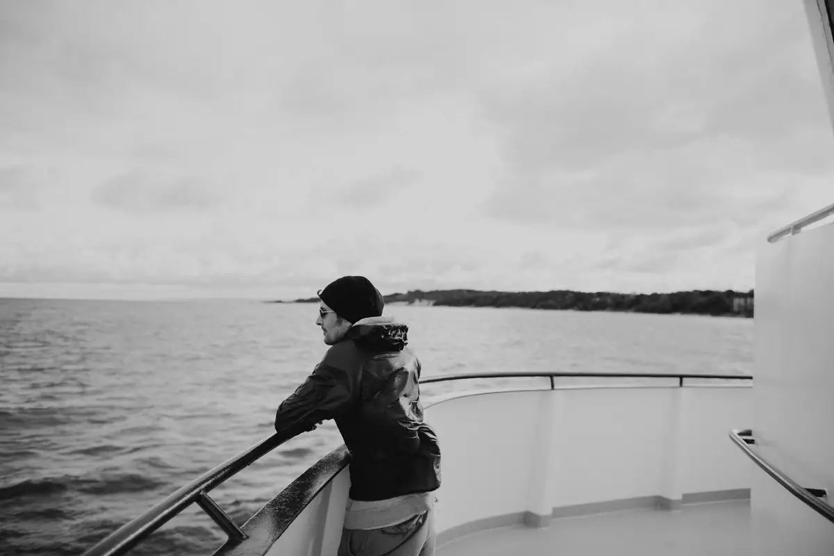 A person leans on a passenger vessel's railing while looking toward a wooded shoreline.