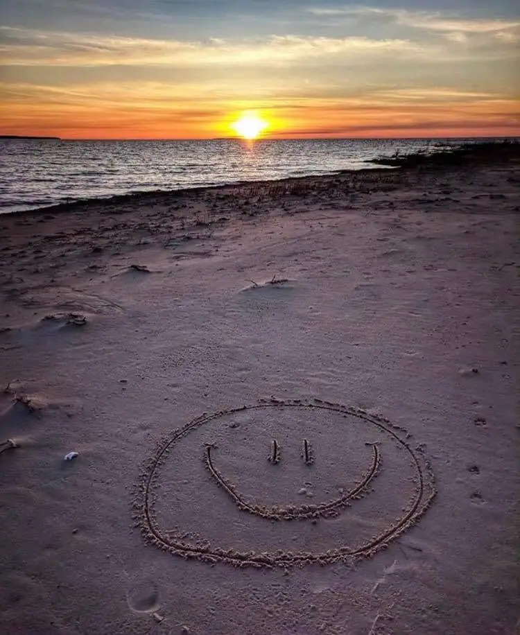 A smiling face drawn in wet sand overlooks a rippling shoreline beneath a vivid sunset.