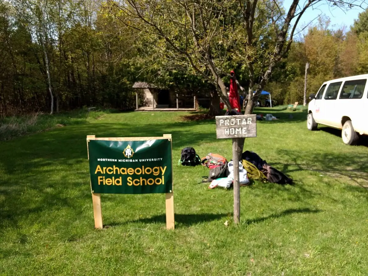 Signs for an archaeology field school and “PROTAR HOME” stand in a grassy, wooded yard beside a white van.