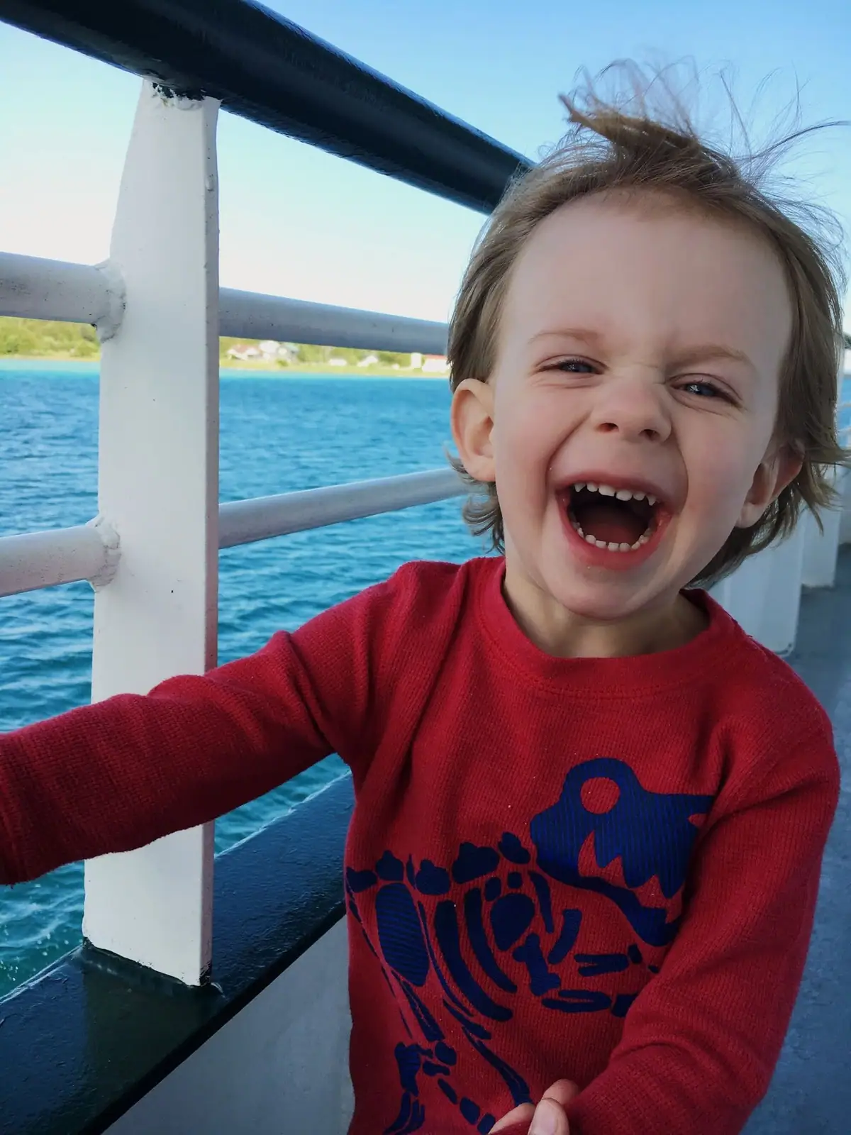 Child laughing beside a white railing on a boat deck, with blue water and a distant shoreline behind.