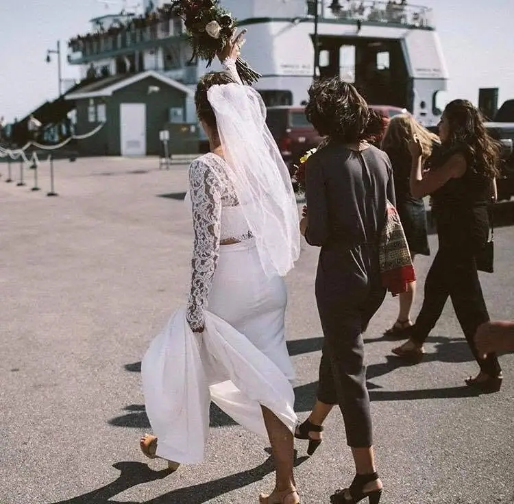 A bride in a white lace dress and veil walks with several people in front of a large ferry.