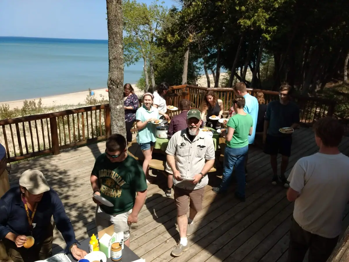 A group eats outdoors on a wooden deck beside a sandy shoreline and tree-covered area.