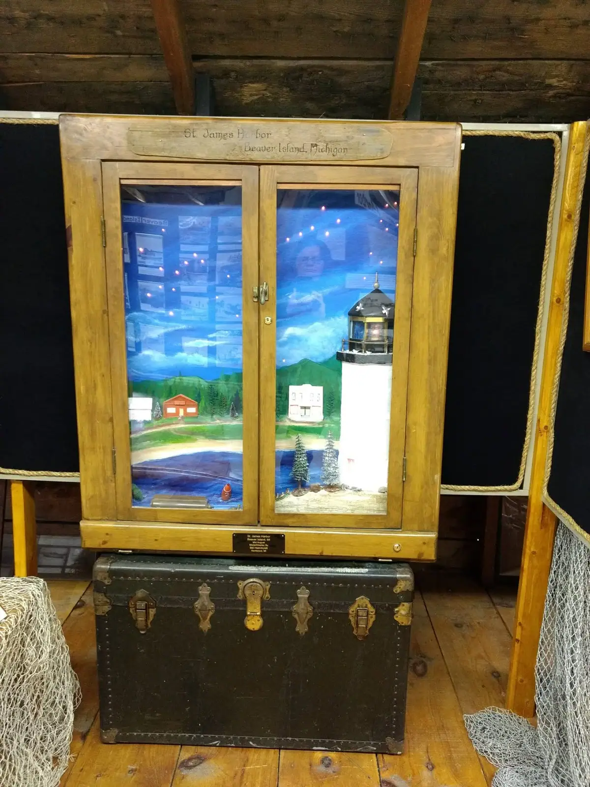 Photograph of a wooden display case holding a painted harbor scene with a lighthouse, buildings, trees, and water.