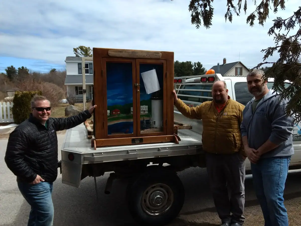 Three men stand beside a truck carrying a wooden glass-front display case with a landscape artwork.