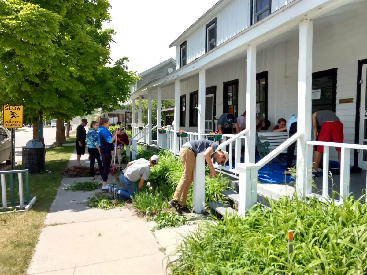 Young people and adults work beside a white wooden building with a porch and a roadside warning sign.