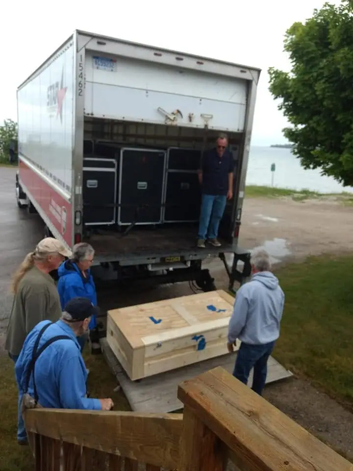 People unload equipment from a box truck beside the water while a large wooden crate rests on a lift.