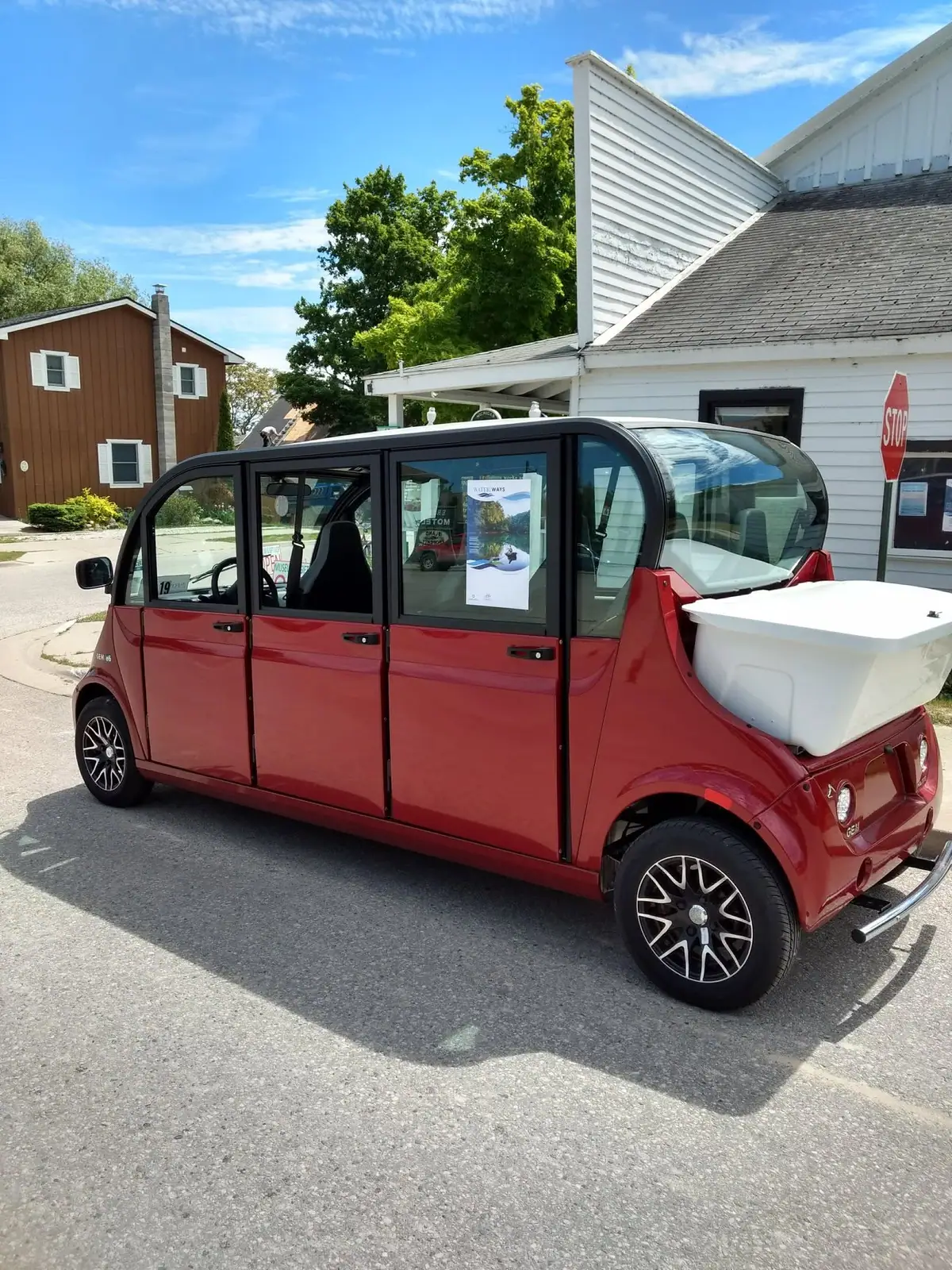 A red electric shuttle with a white rear cargo box is parked beside white and brown buildings.