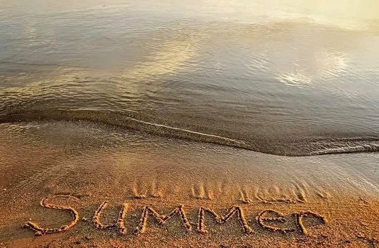 The word “SUMMER” is written in wet sand beside gently rippling water.
