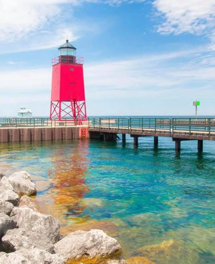 A red lighthouse stands on a pier beside turquoise water, with a white ferry in the distance.
