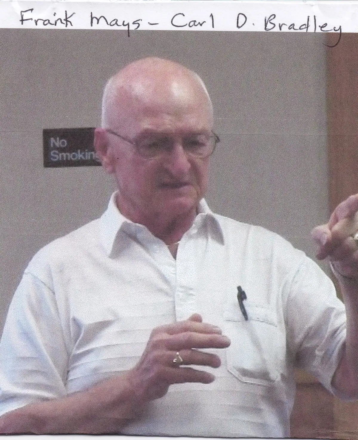 Portrait of a man in a white shirt gesturing beside a No Smoking sign.