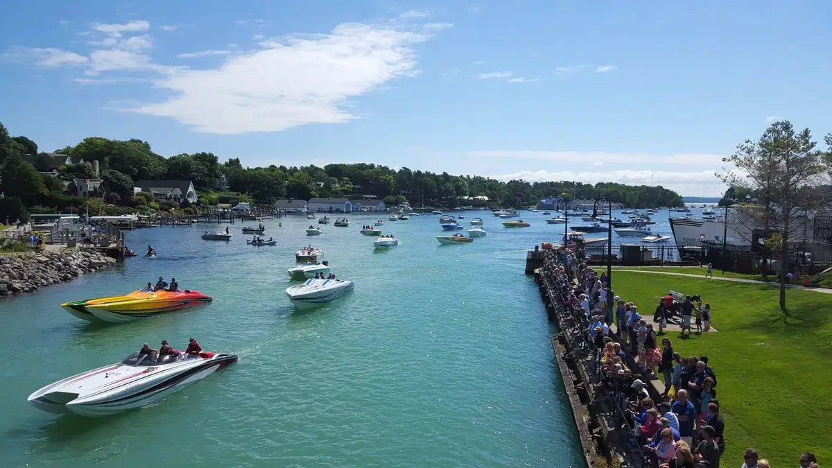Numerous speedboats gather in a waterfront channel as spectators line a pier and grassy shore.