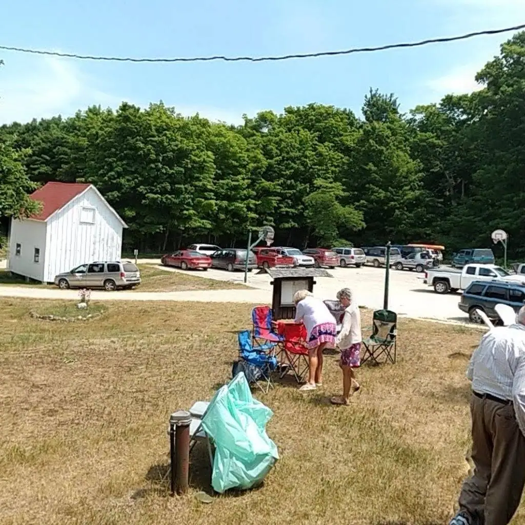 People gather beside folding chairs near a small white outbuilding, parked cars, and a basketball hoop.