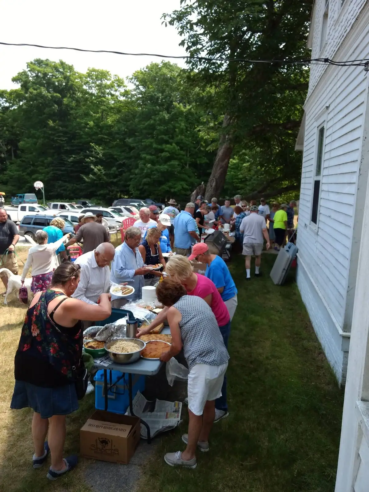 People gather outdoors beside a white building around a table serving food.