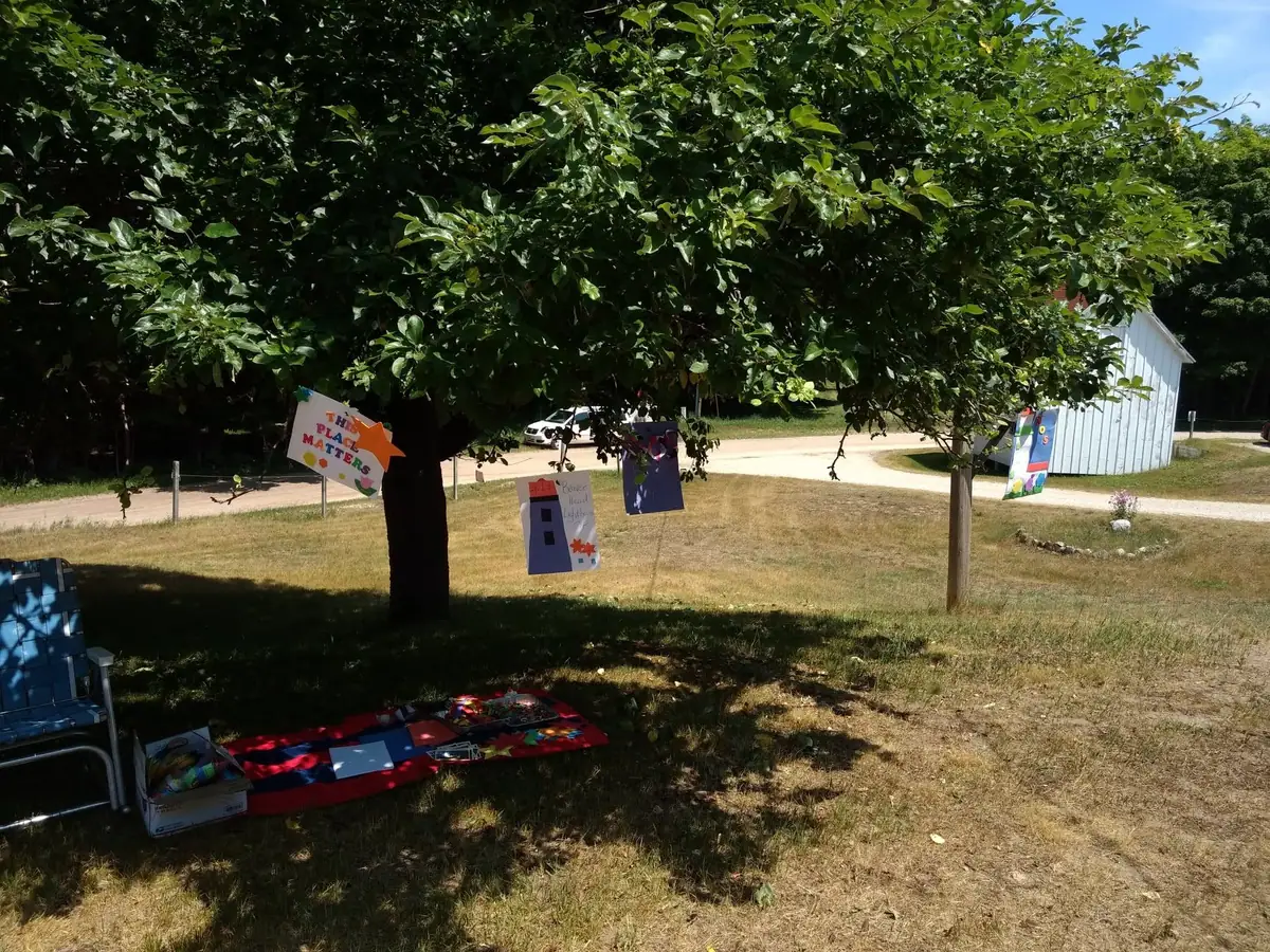 Handmade signs hang from trees beside a chair, blanket, shed, and parked car.