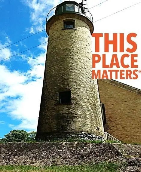A cylindrical stone lighthouse tower rises beside a building beneath a blue, cloud-dotted sky.