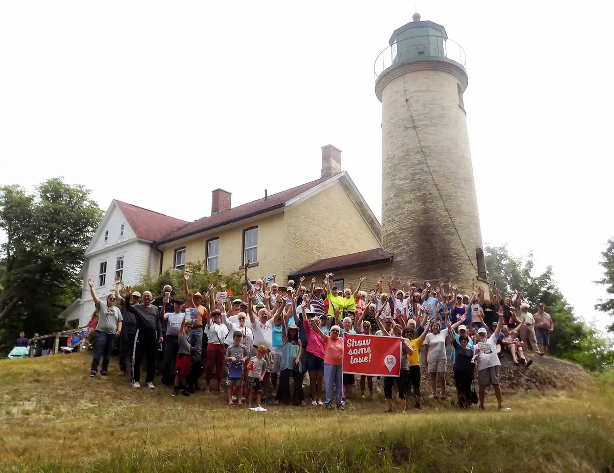 A large group poses with raised arms beside a brick lighthouse and keeper's house, holding a red banner.